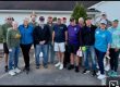 Group of about 16 adults posing together in a parking lot in front of a building, many wearing casual shirts and caps, smiling for the photo.