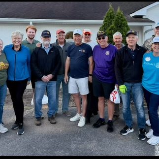 Group of about 16 adults posing together in a parking lot in front of a building, many wearing casual shirts and caps, smiling for the photo.