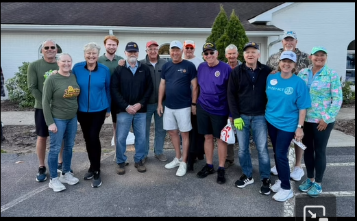 Group of about 16 adults posing together in a parking lot in front of a building, many wearing casual shirts and caps, smiling for the photo.