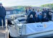 Group of adults on a docked white pontoon boat with a Rotary Impact Club of Clayton banner; a man with a cane stands on the left dock under a sunny sky.
