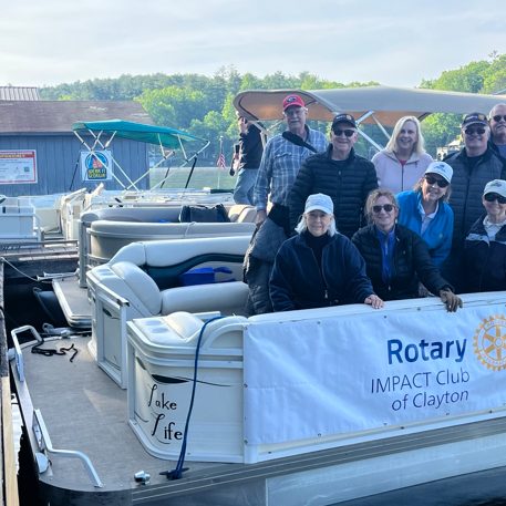 Group of adults on a docked white pontoon boat with a Rotary Impact Club of Clayton banner; a man with a cane stands on the left dock under a sunny sky.