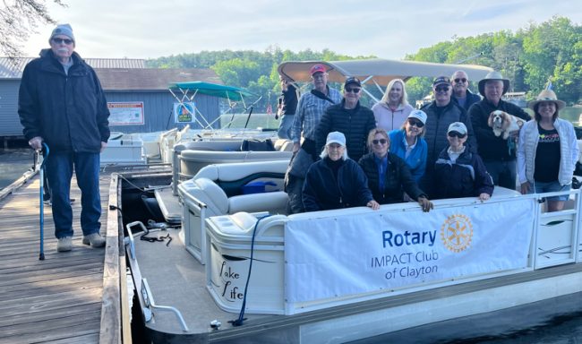 Group of adults on a docked white pontoon boat with a Rotary Impact Club of Clayton banner; a man with a cane stands on the left dock under a sunny sky.