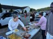 Volunteers serve meals from aluminum trays at a community food distribution in a parking lot outside a church, with cars lining up behind them.