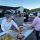 Volunteers serve meals from aluminum trays at a community food distribution in a parking lot outside a church, with cars lining up behind them.