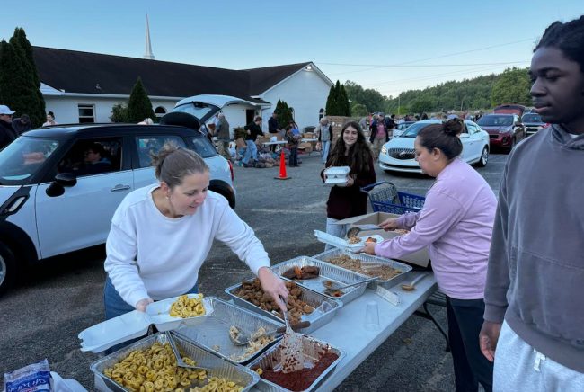 Volunteers serve meals from aluminum trays at a community food distribution in a parking lot outside a church, with cars lining up behind them.
