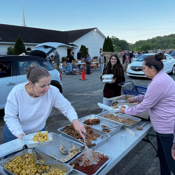 Volunteers serve meals from aluminum trays at a community food distribution in a parking lot outside a church, with cars lining up behind them.