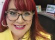 Woman with red hair and glasses, smiling at the camera in a yellow blouse.