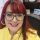 Woman with red hair and glasses, smiling at the camera in a yellow blouse.