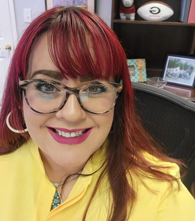 Woman with red hair and glasses, smiling at the camera in a yellow blouse.