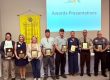 Nine award recipients pose with plaques at an awards presentation, with a Rotary banner and an 'Awards Presentations' slide behind them.