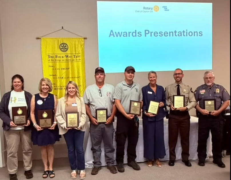 Nine award recipients pose with plaques at an awards presentation, with a Rotary banner and an 'Awards Presentations' slide behind them.