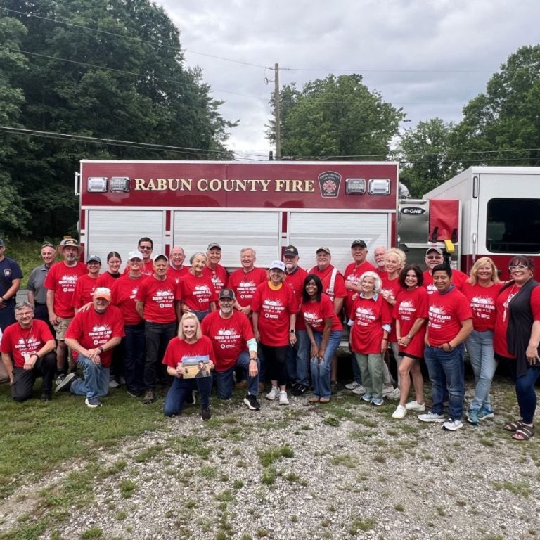 Group of people in red shirts posing in front of a Rabun County Fire engine.