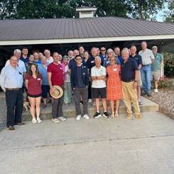 Group of about 25 adults posing together outdoors in front of a building with a covered porch and gravel ground surface.