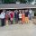 Group of about 25 adults posing together outdoors in front of a building with a covered porch and gravel ground surface.