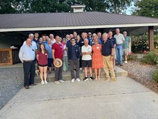 Group of about 25 adults posing together outdoors in front of a building with a covered porch and gravel ground surface.